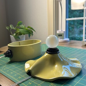 May include: A two-piece chartreuse ceramic lamp featuring a wavy, flared base and a separate oval-shaped housing. The lamp includes a black socket with a round, frosted white light bulb and a clear power cord extending from the base.
