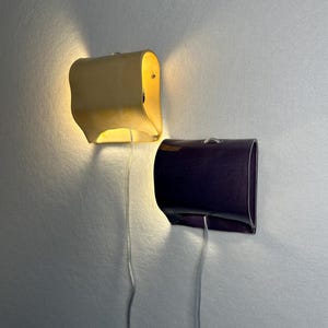 May include: Two wall-mounted sconces with curved, rectangular shades. One sconce is a light cream color and emits a warm, glowing light from the top and bottom. The other sconce is a dark purple color and is unlit. Both have thin, clear power cords.