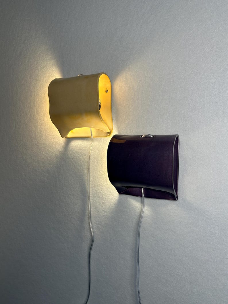 May include: Two wall-mounted sconces with curved, rectangular shades. One sconce is a light cream color and emits a warm, glowing light from the top and bottom. The other sconce is a dark purple color and is unlit. Both have thin, clear power cords.
