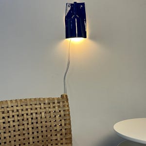 Puede incluir: Un aplique de pared de cer&aacute;mica azul oscuro brillante con forma cil&iacute;ndrica y borde superior plano. La l&aacute;mpara emite una luz c&aacute;lida hacia abajo y est&aacute; conectada a un cable de alimentaci&oacute;n blanco fino que cuelga verticalmente.