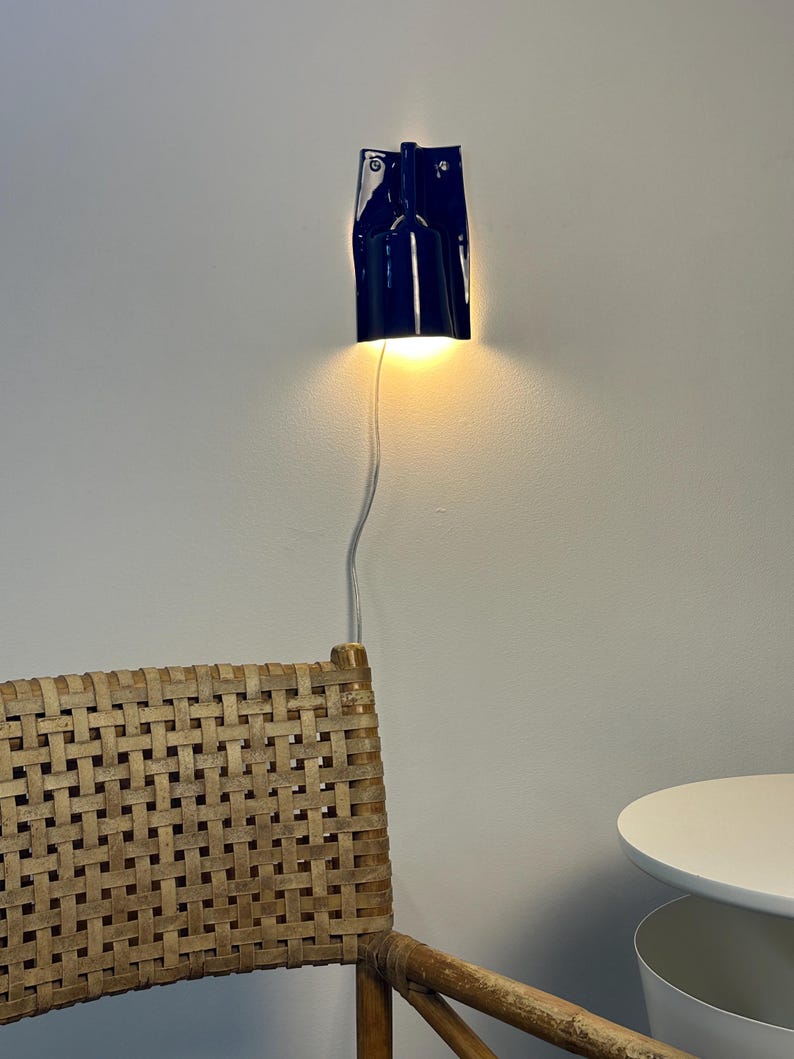 Puede incluir: Un aplique de pared de cer&aacute;mica azul oscuro brillante con forma cil&iacute;ndrica y borde superior plano. La l&aacute;mpara emite una luz c&aacute;lida hacia abajo y est&aacute; conectada a un cable de alimentaci&oacute;n blanco fino que cuelga verticalmente.