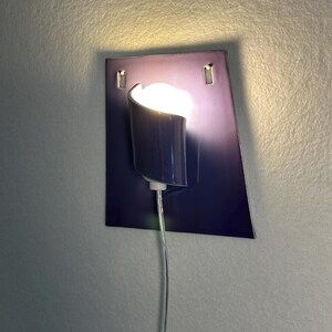 Pode incluir: Uma arandela de parede moderna com uma placa traseira retangular azul escuro. Uma lumin&aacute;ria cil&iacute;ndrica com uma parte superior curva &eacute; montada na placa, emitindo um brilho quente. Um cabo transparente se estende para baixo.