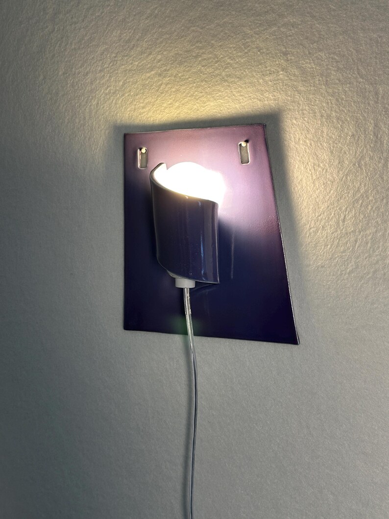 Pode incluir: Uma arandela de parede moderna com uma placa traseira retangular azul escuro. Uma lumin&aacute;ria cil&iacute;ndrica com uma parte superior curva &eacute; montada na placa, emitindo um brilho quente. Um cabo transparente se estende para baixo.