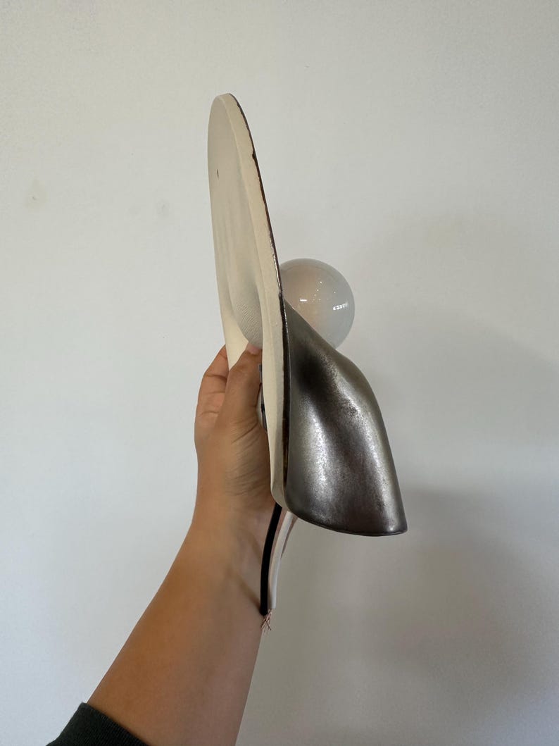 May include: A wall-mounted light fixture featuring a large, flat, off-white circular backplate and a smaller, metallic silver, curved shade. A clear, spherical glass light bulb is positioned between the backplate and the silver shade.