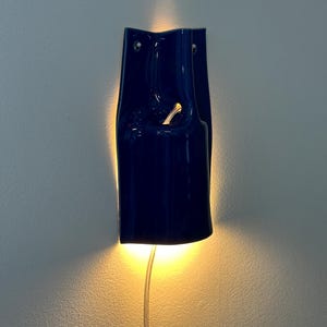 Puede incluir: Un aplique de pared de cer&aacute;mica azul oscuro con acabado brillante, de forma curva y alargada, que emite una luz c&aacute;lida desde la parte posterior y a trav&eacute;s de una abertura central. Un cable de alimentaci&oacute;n fino y transparente sale de la base.