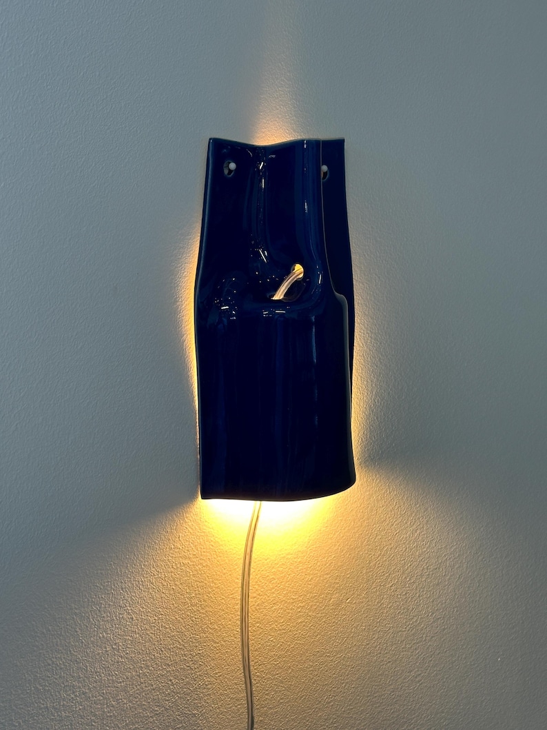 Puede incluir: Un aplique de pared de cer&aacute;mica azul oscuro con acabado brillante, de forma curva y alargada, que emite una luz c&aacute;lida desde la parte posterior y a trav&eacute;s de una abertura central. Un cable de alimentaci&oacute;n fino y transparente sale de la base.