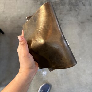 May include: A rectangular, metallic bronze-coloured object with a smooth, curved surface and a slightly distressed, darkened patina. The item features a solid, heavy appearance with rounded edges and a subtle, uneven texture across its front face.