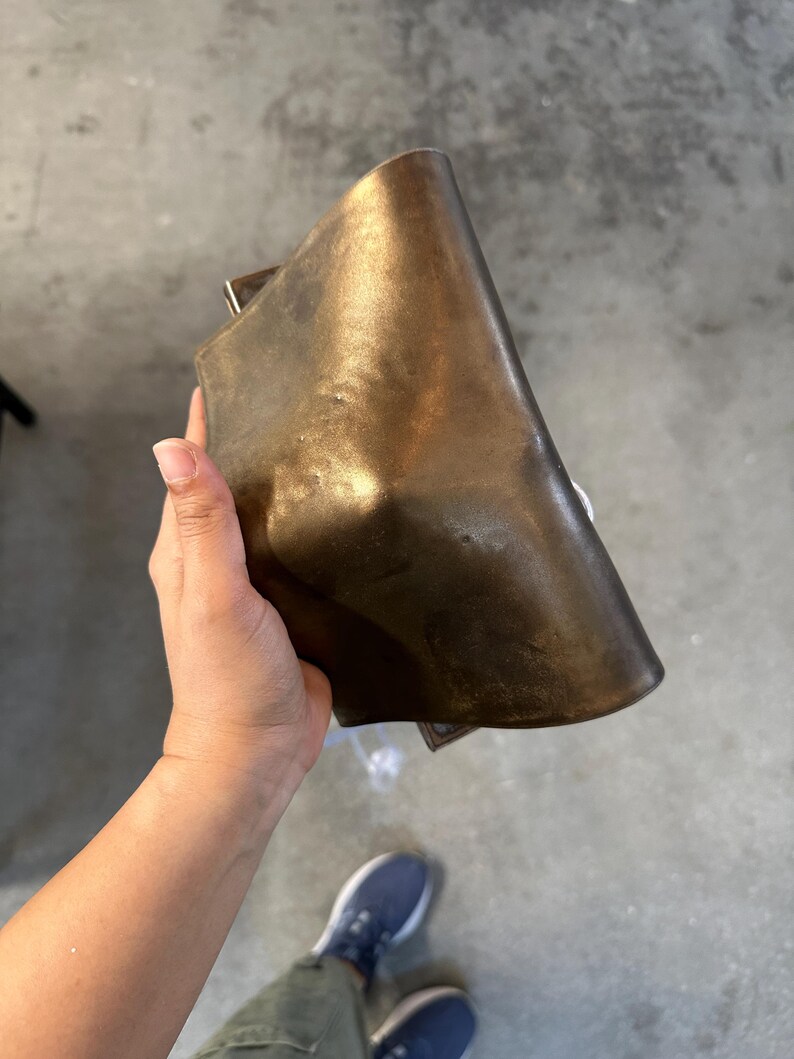May include: A rectangular, metallic bronze-coloured object with a smooth, curved surface and a slightly distressed, darkened patina. The item features a solid, heavy appearance with rounded edges and a subtle, uneven texture across its front face.