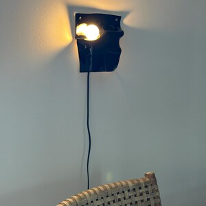 Puede incluir: Un aplique de pared negro con una forma irregular y plegada que emite una luz c&aacute;lida y brillante desde una sola bombilla. La l&aacute;mpara presenta una superficie oscura y lisa y est&aacute; conectada a un fino cable de alimentaci&oacute;n negro que cuelga.