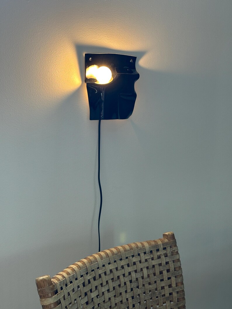 Puede incluir: Un aplique de pared negro con una forma irregular y plegada que emite una luz c&aacute;lida y brillante desde una sola bombilla. La l&aacute;mpara presenta una superficie oscura y lisa y est&aacute; conectada a un fino cable de alimentaci&oacute;n negro que cuelga.