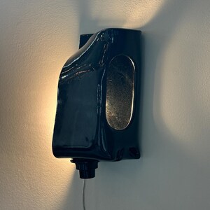 May include: A dark navy blue ceramic wall sconce with a glossy finish and an elongated oval cutout on the side. The fixture is mounted to a wall and emits a warm, soft glow from behind its curved, sculptural form, featuring a small black base component.