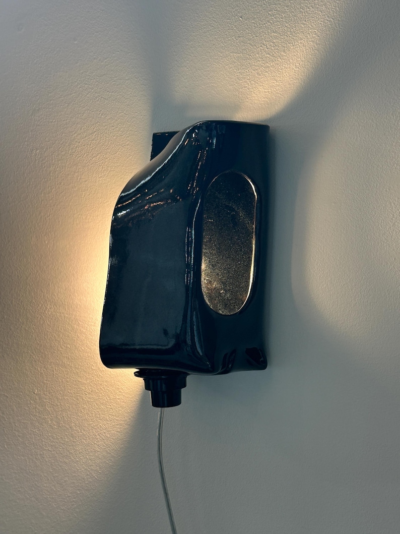 May include: A dark navy blue ceramic wall sconce with a glossy finish and an elongated oval cutout on the side. The fixture is mounted to a wall and emits a warm, soft glow from behind its curved, sculptural form, featuring a small black base component.