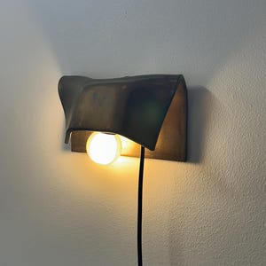 Pode incluir: Um candeeiro de parede com um abajur de metal escuro e curvo com acabamento mate. Uma l&acirc;mpada redonda e brilhante est&aacute; posicionada sob o abajur, emitindo uma luz quente. Um fio de alimenta&ccedil;&atilde;o preto fino desce a partir da lumin&aacute;ria.