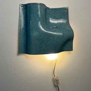 May include: A wall-mounted light fixture with a glossy, teal-blue ceramic shade featuring a dark speckled pattern. The shade has a curved, wave-like form with two mounting holes at the top corners and a small metal loop, emitting a warm light.