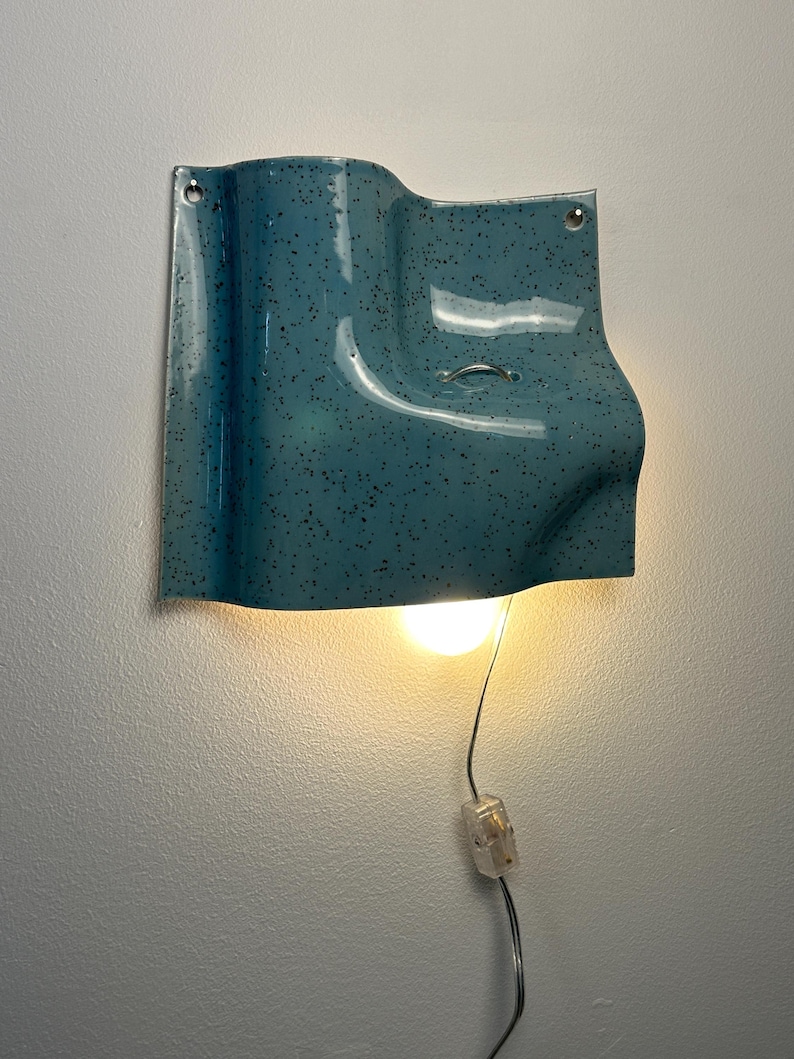 May include: A wall-mounted light fixture with a glossy, teal-blue ceramic shade featuring a dark speckled pattern. The shade has a curved, wave-like form with two mounting holes at the top corners and a small metal loop, emitting a warm light.
