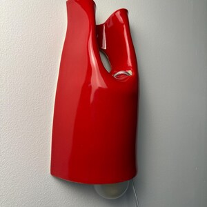 May include: A glossy red ceramic wall sconce with a curved, abstract silhouette featuring a hollow cutout section. A single light bulb is visible at the bottom of the fixture, and a thin, clear electrical cord extends downward from the base.