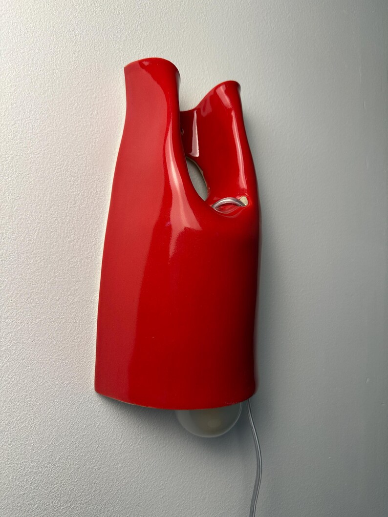 May include: A glossy red ceramic wall sconce with a curved, abstract silhouette featuring a hollow cutout section. A single light bulb is visible at the bottom of the fixture, and a thin, clear electrical cord extends downward from the base.