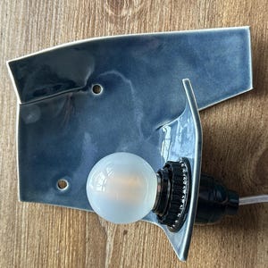 May include: A blue ceramic wall sconce with a bulb. The sconce has a unique, asymmetrical shape with a glossy finish. The bulb is round and emits a soft glow. The sconce is mounted on a wooden surface.