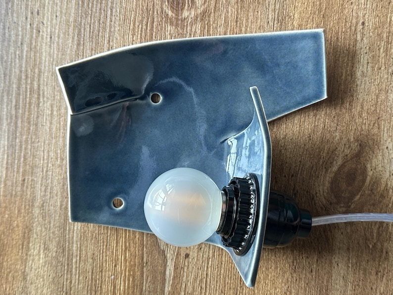 May include: A blue ceramic wall sconce with a bulb. The sconce has a unique, asymmetrical shape with a glossy finish. The bulb is round and emits a soft glow. The sconce is mounted on a wooden surface.