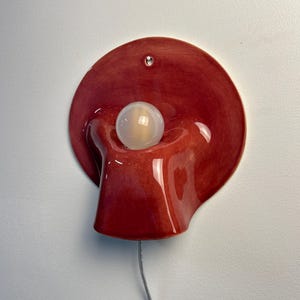 May include: A wall-mounted ceramic lamp featuring a glossy, deep red, organic, fluid-shaped base with a central circular opening. A single, spherical, frosted white light bulb is positioned within the centre, and a thin, clear power cord extends downwards.