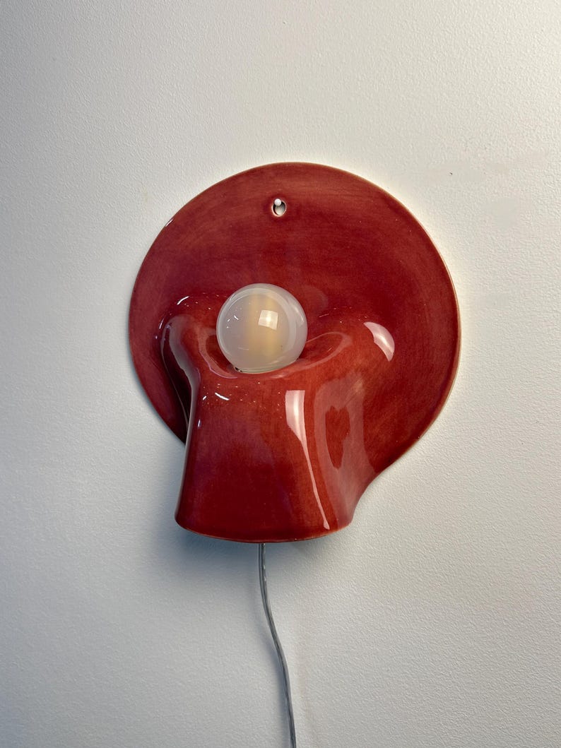 May include: A wall-mounted ceramic lamp featuring a glossy, deep red, organic, fluid-shaped base with a central circular opening. A single, spherical, frosted white light bulb is positioned within the centre, and a thin, clear power cord extends downwards.