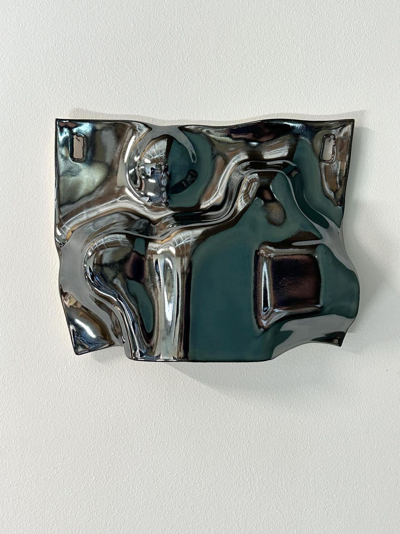 Pu&ograve; includere: Una placca da parete rettangolare in ceramica con una smaltatura argentata metallizzata altamente riflettente. La superficie presenta una texture irregolare, ondulata e simile a un liquido con curve e pieghe profonde. Due piccoli fori rettangolari sono posizionati in alto.