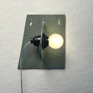 May include: A wall-mounted light fixture featuring a trapezoidal, speckled blue-green ceramic backplate with two mounting holes. A black socket holds a glowing, round white light bulb, which is positioned in front of a circular ceramic disc.