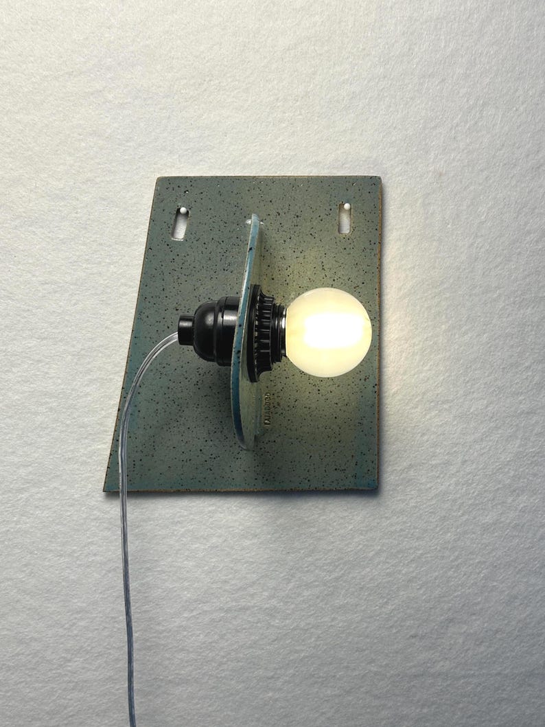 May include: A wall-mounted light fixture featuring a trapezoidal, speckled blue-green ceramic backplate with two mounting holes. A black socket holds a glowing, round white light bulb, which is positioned in front of a circular ceramic disc.
