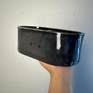 May include: A rectangular, black ceramic planter with a glossy finish. The planter has rounded corners and a smooth surface. The dark colour and simple shape make it suitable for various plants and decor styles.