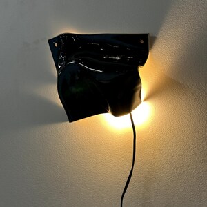 Puede incluir: Un aplique de pared con una pantalla de cer&aacute;mica negra brillante de forma irregular y aspecto fluido y drapeado. La l&aacute;mpara emite una luz c&aacute;lida y dorada desde detr&aacute;s de la pantalla, proyectando un suave resplandor en la pared, con un cable negro.