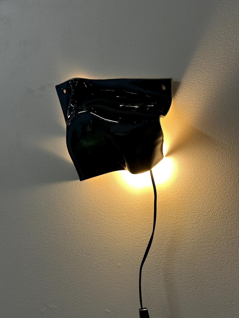 Puede incluir: Un aplique de pared con una pantalla de cer&aacute;mica negra brillante de forma irregular y aspecto fluido y drapeado. La l&aacute;mpara emite una luz c&aacute;lida y dorada desde detr&aacute;s de la pantalla, proyectando un suave resplandor en la pared, con un cable negro.