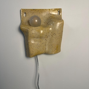 May include: A wall-mounted light fixture featuring a beige ceramic base with a dark brown speckled pattern. The base has an irregular, undulating shape with two rectangular cutouts at the top and a single round, frosted glass light bulb.