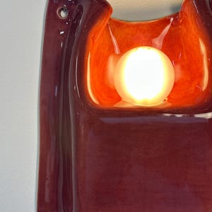 Puede incluir: Un aplique de pared de cer&aacute;mica con un exterior brillante de color burdeos intenso y un cuenco interior de color naranja vibrante y luminoso. Una bombilla esf&eacute;rica est&aacute; colocada en el centro de la forma cer&aacute;mica curva y org&aacute;nica.