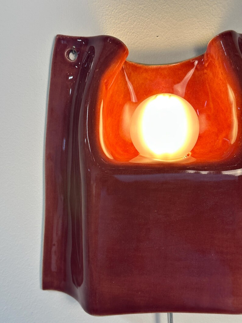 Puede incluir: Un aplique de pared de cer&aacute;mica con un exterior brillante de color burdeos intenso y un cuenco interior de color naranja vibrante y luminoso. Una bombilla esf&eacute;rica est&aacute; colocada en el centro de la forma cer&aacute;mica curva y org&aacute;nica.
