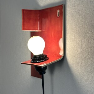 Pode incluir: Uma moderna arandela de parede de cer&acirc;mica vermelha com um design curvo. Possui uma pequena prateleira que suporta uma l&acirc;mpada redonda e iluminada com uma base preta. A arandela &eacute; montada em uma parede cinza e tem um cabo de alimenta&ccedil;&atilde;o preto.