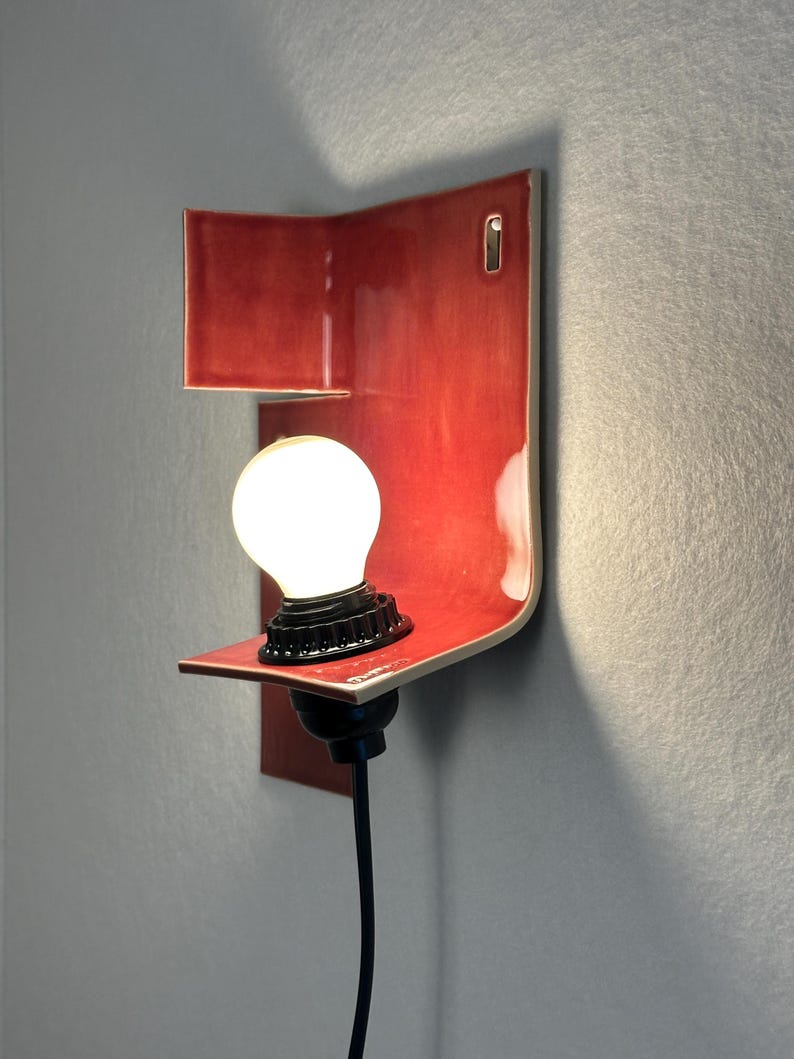 Pode incluir: Uma moderna arandela de parede de cer&acirc;mica vermelha com um design curvo. Possui uma pequena prateleira que suporta uma l&acirc;mpada redonda e iluminada com uma base preta. A arandela &eacute; montada em uma parede cinza e tem um cabo de alimenta&ccedil;&atilde;o preto.