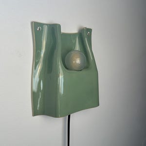 May include: A sage green ceramic wall light with a glossy finish and a wavy, organic silhouette. The fixture features a central cutout holding a single frosted glass globe light bulb. A thin black power cord extends from the bottom of the unit.