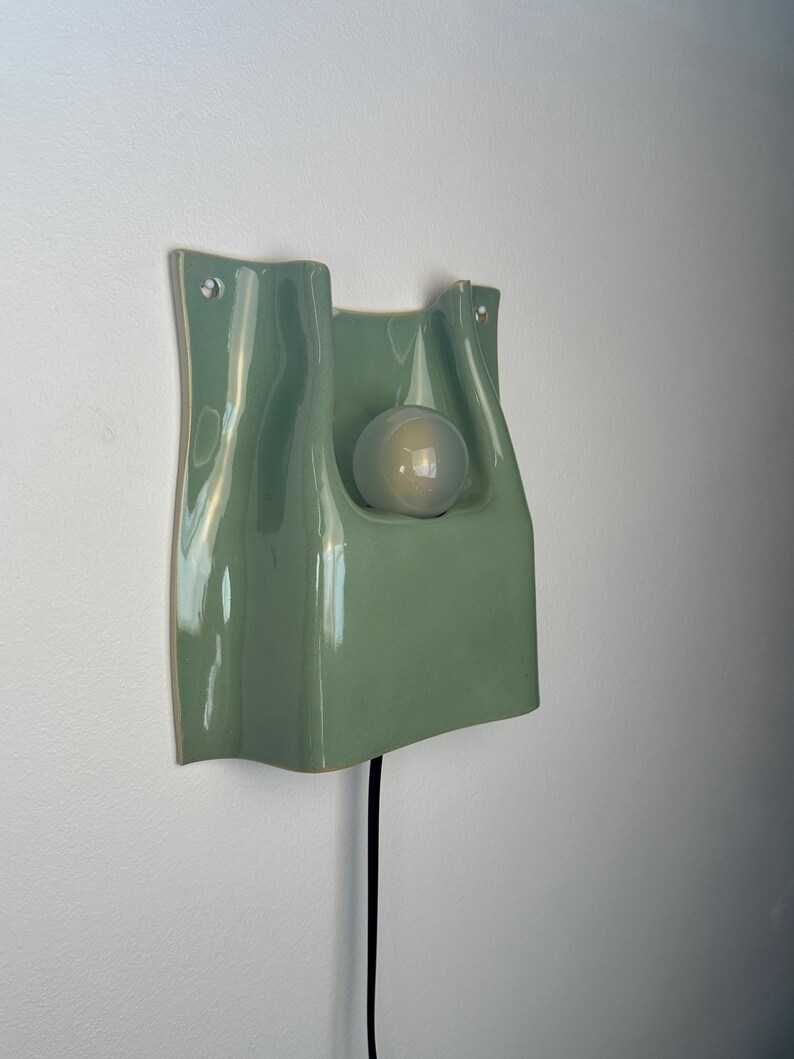 May include: A sage green ceramic wall light with a glossy finish and a wavy, organic silhouette. The fixture features a central cutout holding a single frosted glass globe light bulb. A thin black power cord extends from the bottom of the unit.