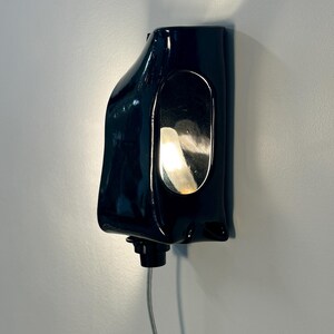 May include: A glossy black ceramic wall sconce with an organic, asymmetrical shape. The fixture features a central oval cutout that reveals a warm, glowing light source from within. A small black knob is located at the bottom, connected to a thin cord.