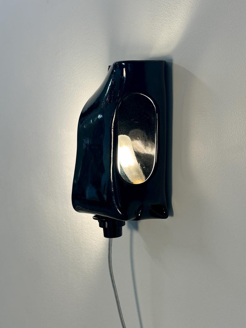 May include: A glossy black ceramic wall sconce with an organic, asymmetrical shape. The fixture features a central oval cutout that reveals a warm, glowing light source from within. A small black knob is located at the bottom, connected to a thin cord.