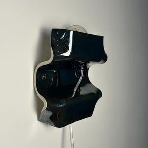 May include: A wall-mounted lamp featuring a glossy black, undulating ceramic form with a white interior edge. The fixture includes a central black socket holding a clear light bulb and a transparent electrical cord extending from the bottom.