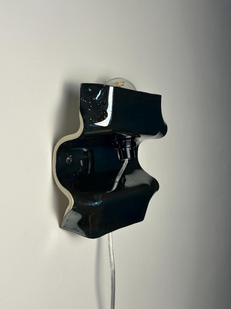 May include: A wall-mounted lamp featuring a glossy black, undulating ceramic form with a white interior edge. The fixture includes a central black socket holding a clear light bulb and a transparent electrical cord extending from the bottom.