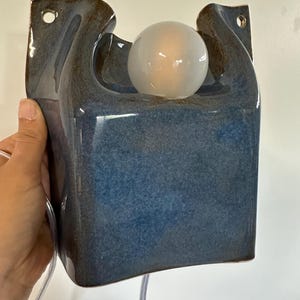 May include: A wall-mounted ceramic lamp featuring a glossy blue glaze with dark brown edges. The rectangular body has a curved top section holding a single round, frosted white light bulb. Two circular holes are positioned at the top corners.
