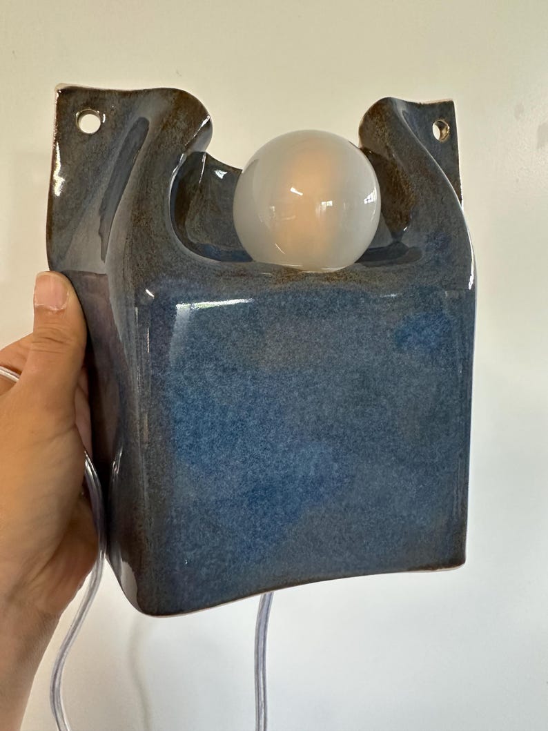 May include: A wall-mounted ceramic lamp featuring a glossy blue glaze with dark brown edges. The rectangular body has a curved top section holding a single round, frosted white light bulb. Two circular holes are positioned at the top corners.