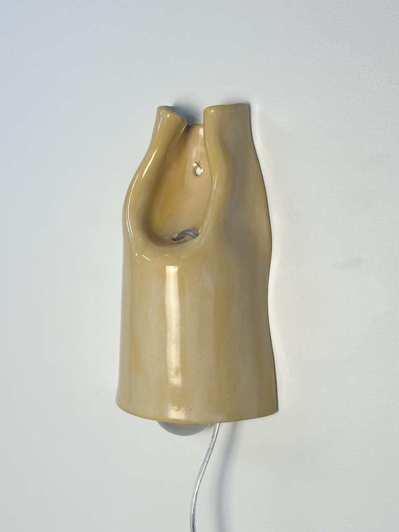Pu&ograve; includere: Una lampada da parete in ceramica giallo pallido lucida con una forma organica e ripiegata. L'oggetto presenta un'apertura centrale per una lampadina, un piccolo foro di montaggio circolare nella parte superiore e un sottile cavo elettrico trasparente.