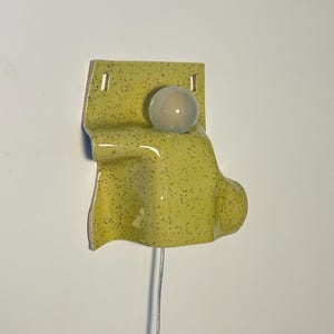 Puede incluir: Una l&aacute;mpara de pared de cer&aacute;mica con un esmalte brillante de color verde amarillento y motas oscuras. El aplique tiene una placa trasera rectangular con dos orificios de montaje y una secci&oacute;n frontal curva y saliente que sostiene una bombilla redonda esmerilada.