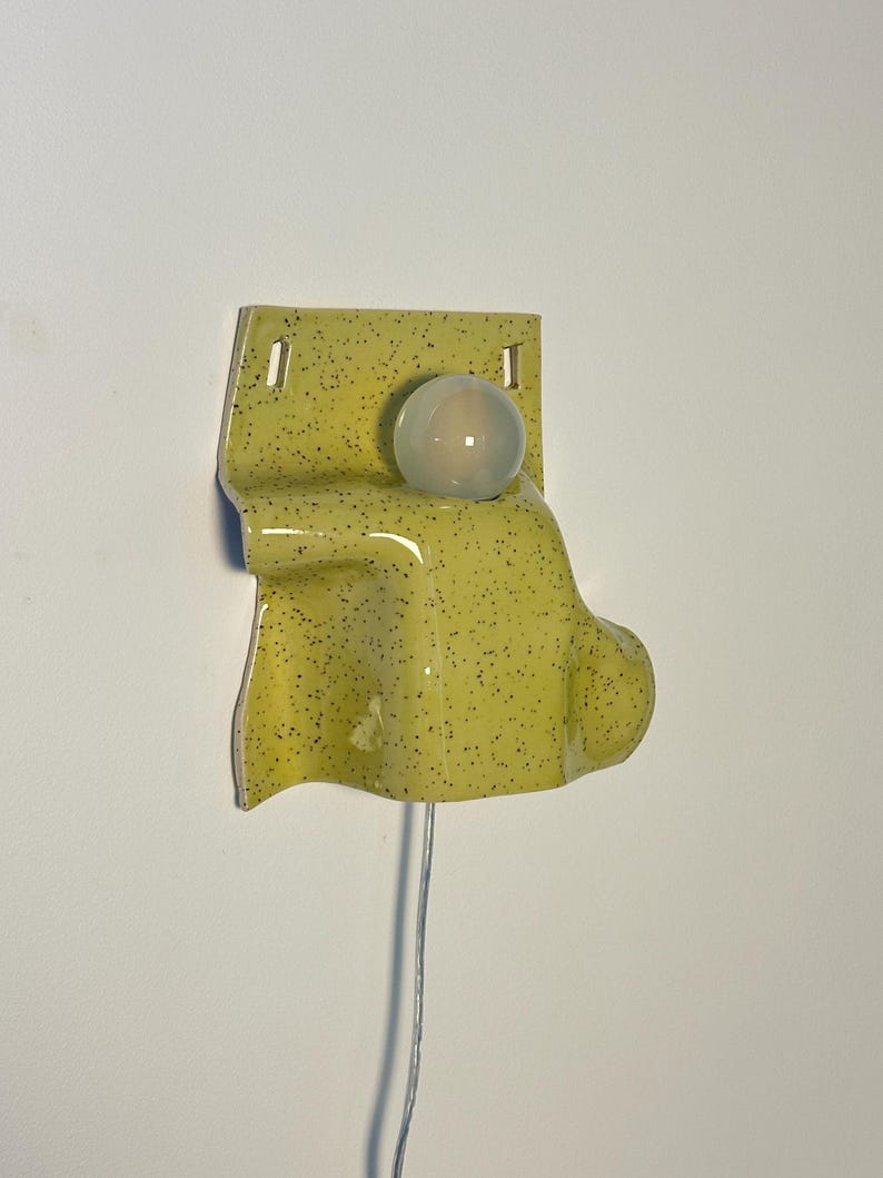 Puede incluir: Una l&aacute;mpara de pared de cer&aacute;mica con un esmalte brillante de color verde amarillento y motas oscuras. El aplique tiene una placa trasera rectangular con dos orificios de montaje y una secci&oacute;n frontal curva y saliente que sostiene una bombilla redonda esmerilada.