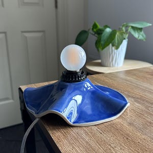 May include: A small, blue ceramic lamp with a unique, ruffled shade design. The lamp features a frosted glass globe bulb and a black base. The lamp is on a wooden surface, with a plant in a white pot in the background.