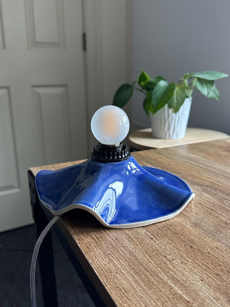 May include: A small, blue ceramic lamp with a unique, ruffled shade design. The lamp features a frosted glass globe bulb and a black base. The lamp is on a wooden surface, with a plant in a white pot in the background.