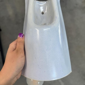K&ouml;nnte beinhalten: Ein hellblauer Wandpflanztopf aus Keramik mit einer glatten, gesprenkelten Oberfl&auml;che. Das Gef&auml;&szlig; verf&uuml;gt &uuml;ber eine breite, ausgestellte &Ouml;ffnung an der Oberseite mit einem kleinen kreisf&ouml;rmigen Loch zur Befestigung und einen abgerundeten Boden.